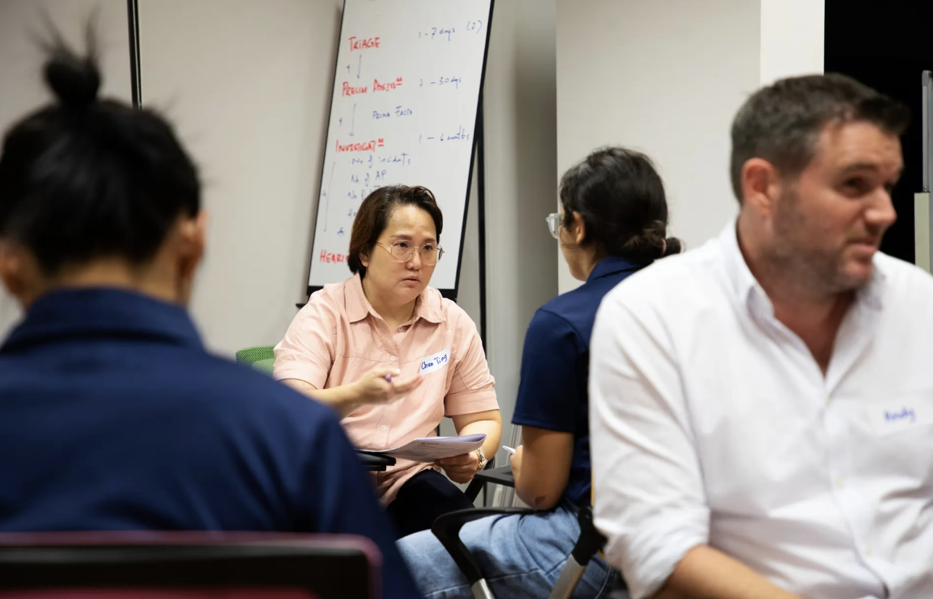 A man and woman engaged in discussion at a table, with a whiteboard displaying notes in the background.